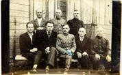 Prie medinio pastato sienos sėdi ant medinio suolo 5 vyrai, už jų 3 stovi. 1946 m., Stakiai (nuotrauka)
